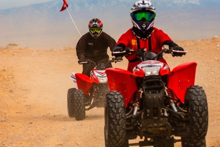 Two people wearing helmets riding red ATVs on a dirt path.