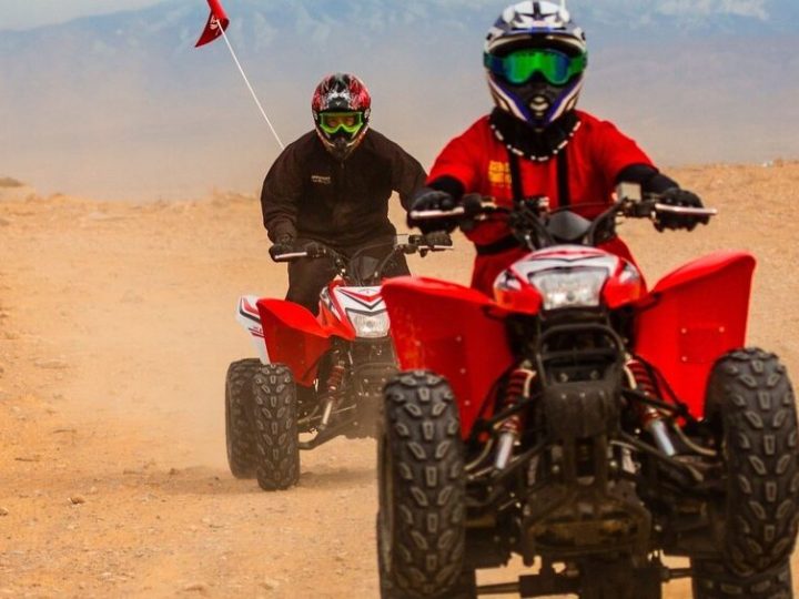 Two people wearing helmets riding red ATVs on a dirt path.