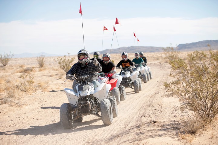 Group of people riding ATVs in desert terrain with red flags on poles.