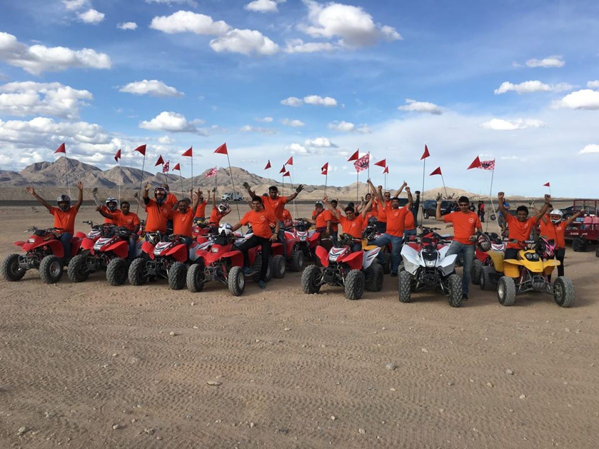 Group of people in orange shirts on quad bikes in a desert with mountains in the background.