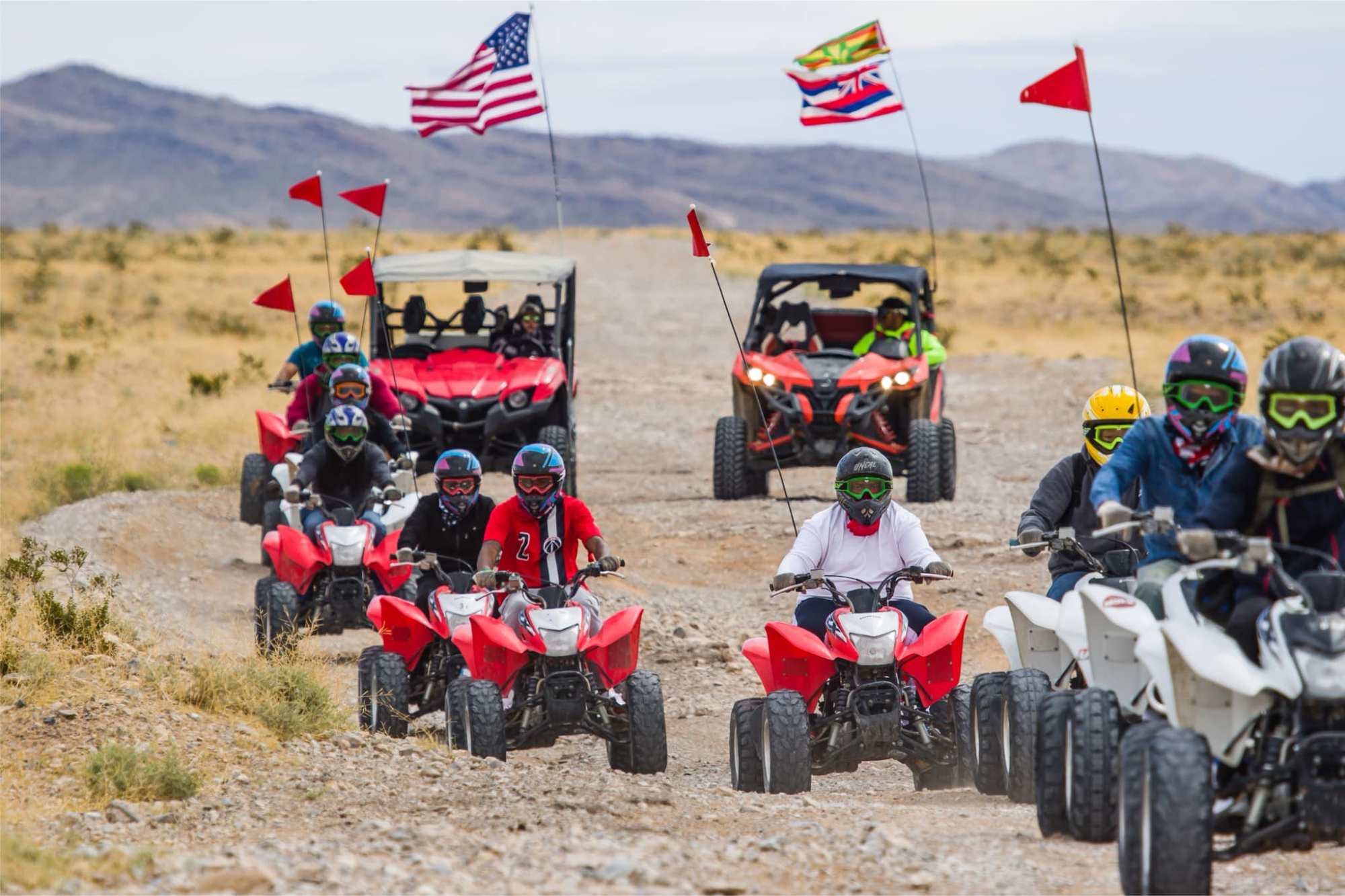 Group of people riding ATVs on dirt road with flags in desert landscape.