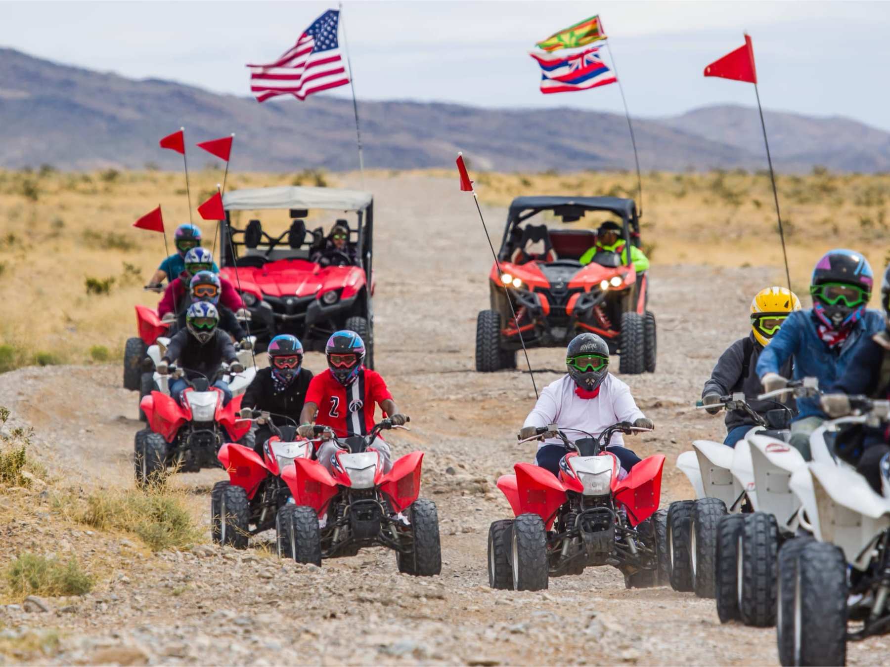 Group of people riding ATVs on dirt road with flags in desert landscape.