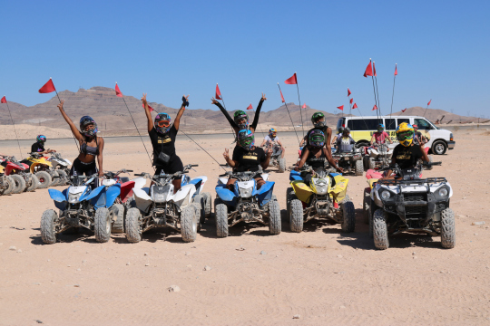 Five people pose on ATVs in a desert with mountains in the background, waving and smiling.