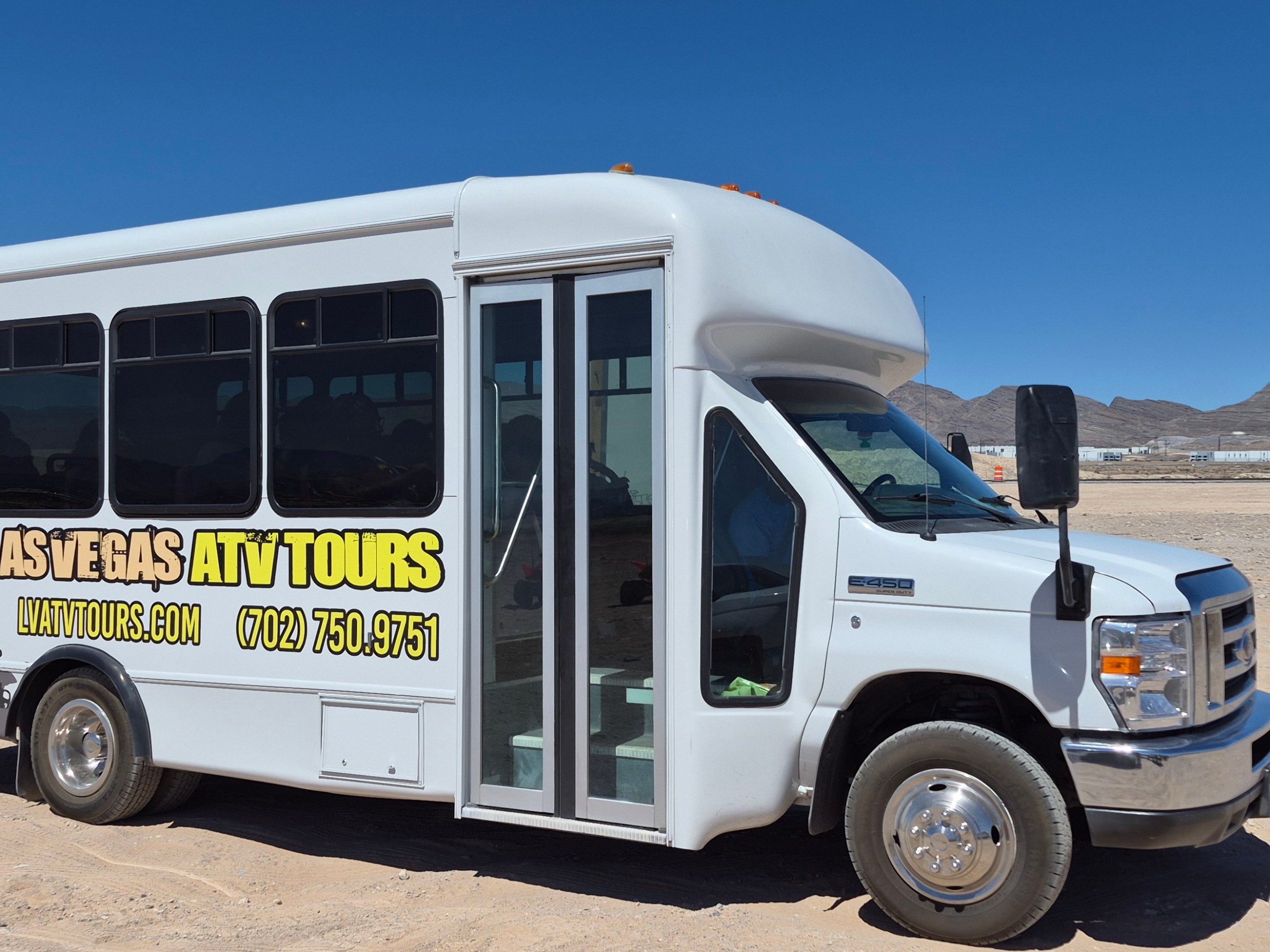 White tour bus parked in desert with mountains in background.