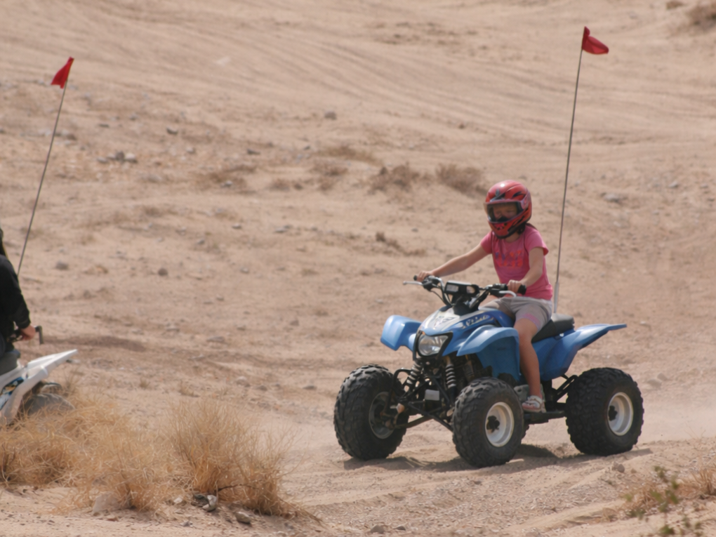 Child in helmet riding blue ATV in desert landscape.